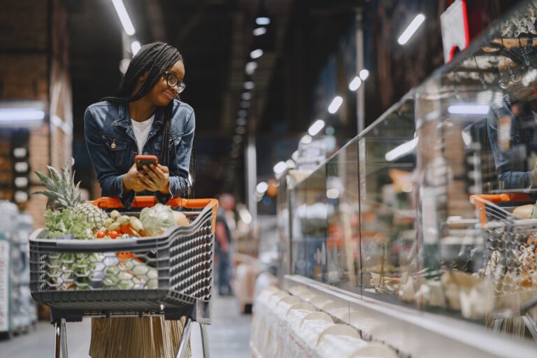 A young woman with afro braids grocery shopping in a supermarket, using a mobile phone.