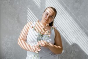 Plus-size woman in tank top holding flowers with sunlight filtering through blinds.