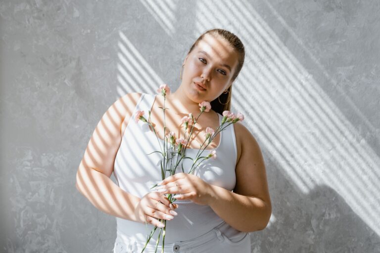 Plus-size woman in tank top holding flowers with sunlight filtering through blinds.